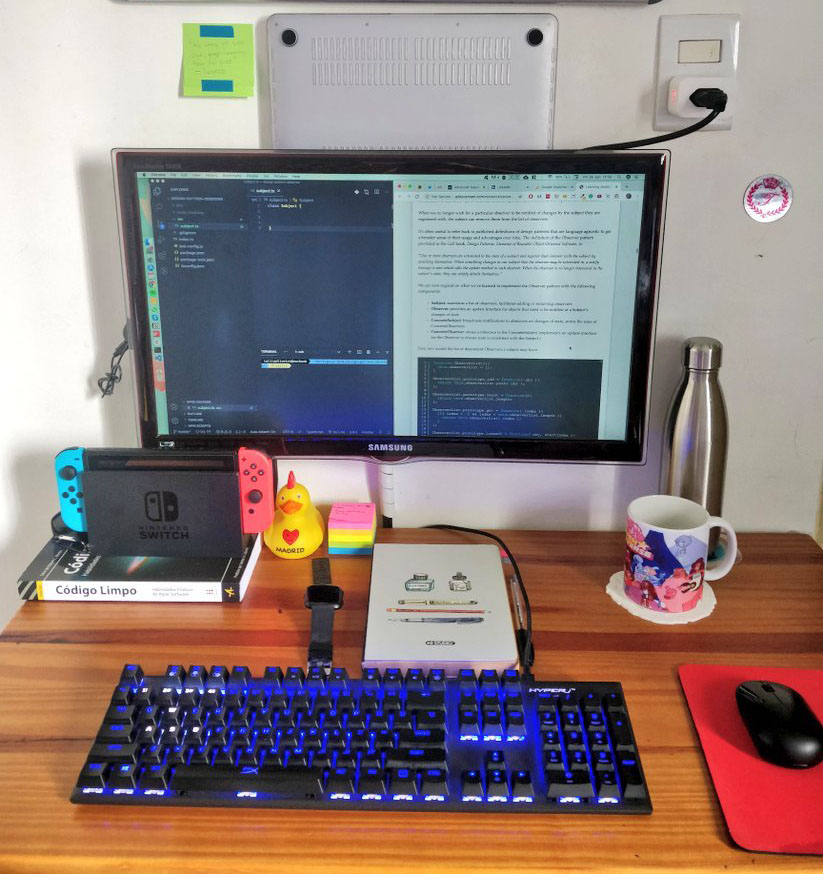 Foto de uma mesa com: teclado mecânico iluminado de azul, um mouse pad vermelho do lado direito com um mouse preto em cima, livro Clean Code, um Nintendo swicth em cima do livro, um pato de borracha ao lado direito dos dois, um relógio e um caderno, uma caneca, uma garrafa de agua e acima da mesa uma televisão na parede como monitor para o laptop que está acima da televisão.