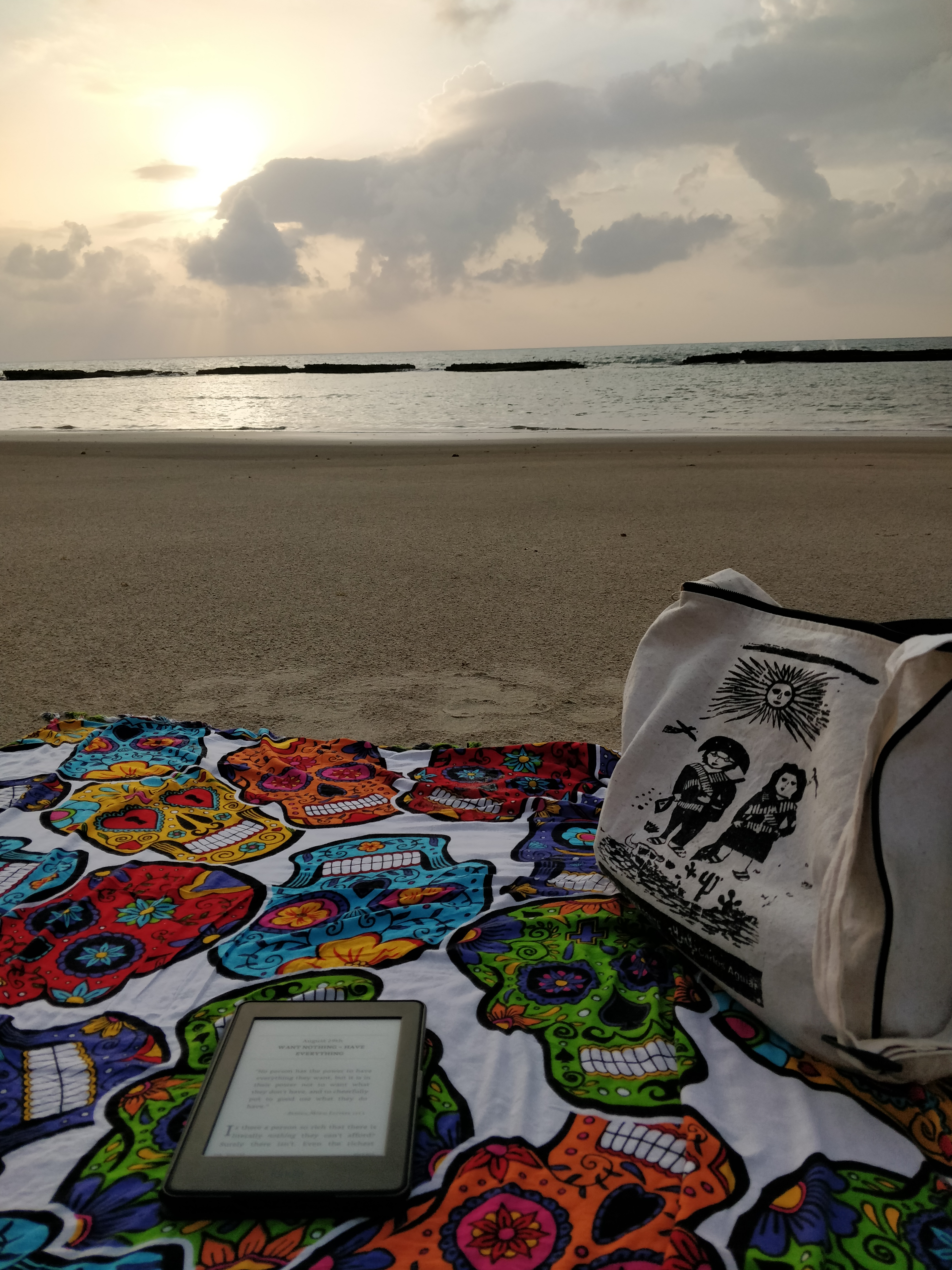 Foto da praia com o sol nascendo ao fundo, e na areia uma canga com um kindle em cima e uma bolsa.